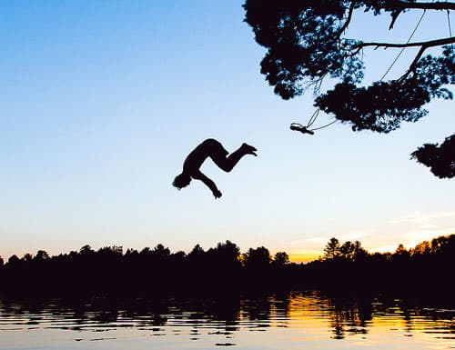 Silhouette of someone in midair as they swing from a tree into a lake.