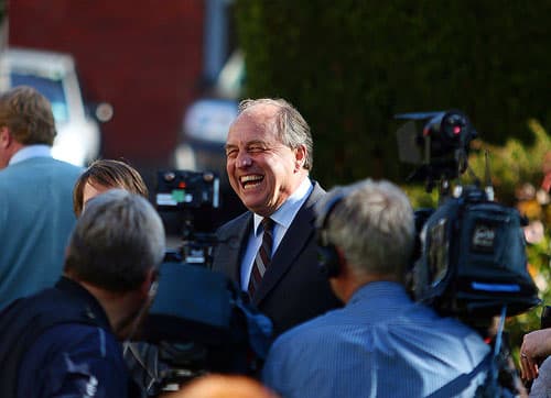 Man in dark suit surrounded by cameras and videographers laughing