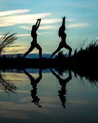 Silhouette of two women stretching at sunrise by beach.