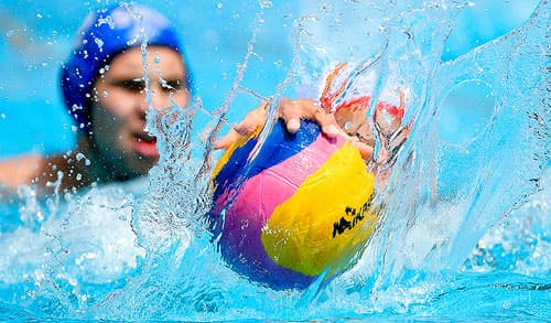 Two sport swimmers reaching for a ball in a pool.