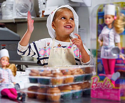 Little girl dressed like a baker surrounded by Barbie dolls in a baker outfit.