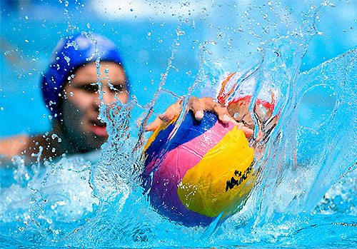 Two sport swimmers reaching for a ball in a pool.