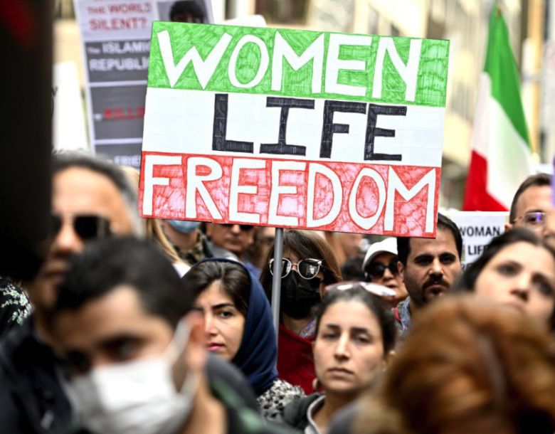An image of protestors in a crowd holding up a sign that says Women, life, freedom in the colours of the Iranian flag.